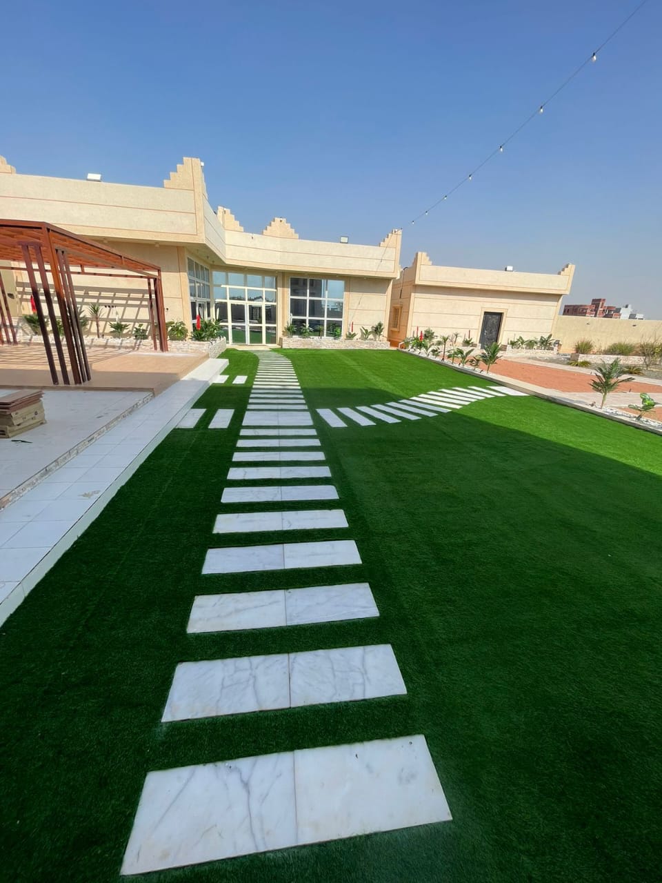 حديقة فيلا في الرياض بمسطح أخضر كثيف وممر رخامي أبيض وبرجولة خشبية من تنفيذ بستان